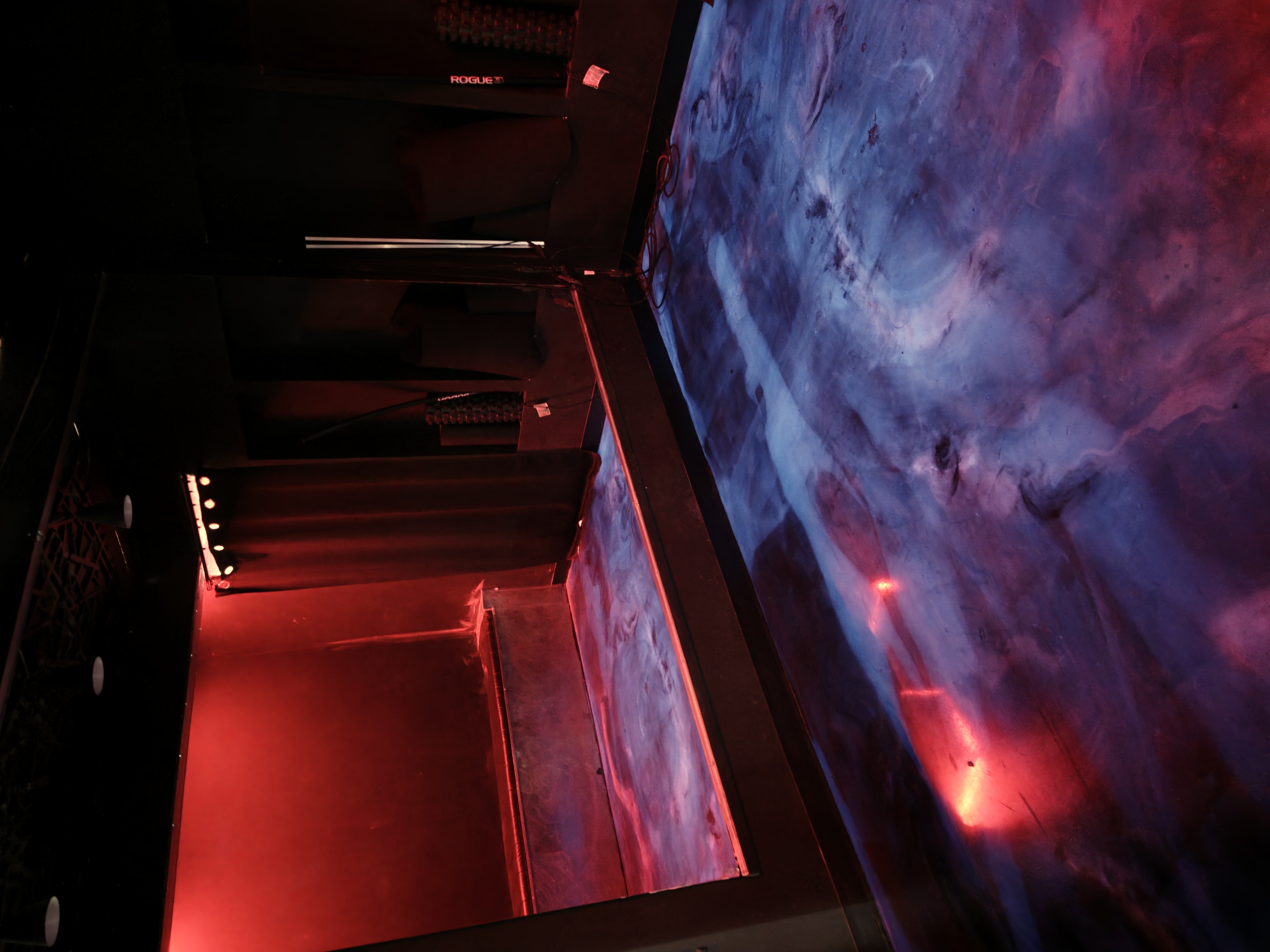 Red-lit posing room with glowing blue floor, mirror, and bench alcove at Iron Arms Gym